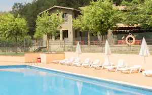 Schwimmbad mit blauen Beckenrand, weißen Liegen und Sonnenschirmen vor einem Steingebäude und Bäumen im Campingbereich.