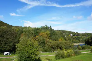 Campingplatz-Panorama mit Wiesen, Bäumen und einem kleinen Wohnwagen am Rand, im Hintergrund ein Fluss und bewaldete Hügel.