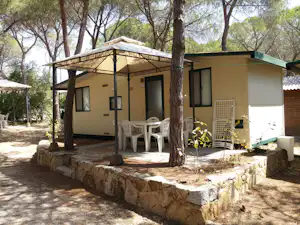 Campingbungalow mit überdachter Terrasse, weißen Stühlen und Tisch unter Bäumen, Steinmauer und Eingangsbereich am Gebäude.