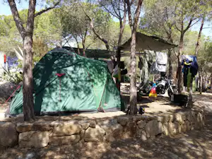 Grünes Campingzelt im Schatten von Bäumen, daneben Motorräder, Wäscheleine und Zeltplanen im Campingbereich.