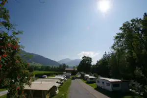 Campingplatz-Allee mit Wohnwagen und Zelten, Rasenstreifen, Bäumen und Bergen im Hintergrund.