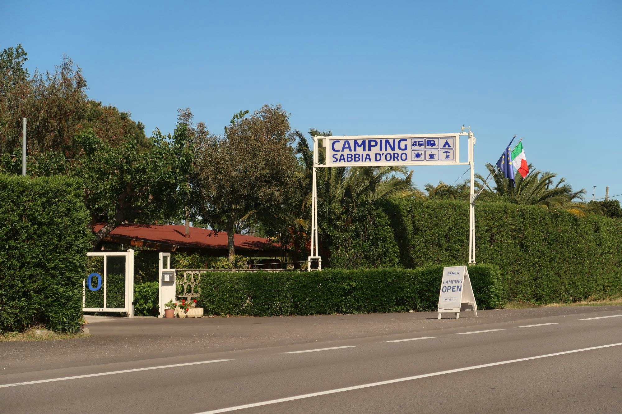 Eingangsschild „Camping Sabbia d’Oro“ am Straßenrand, mit Flaggen, Hecken und Bäumen im Hintergrund.