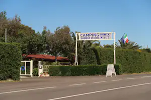 Eingangsschild „Camping Sabbia d’Oro“ am Straßenrand, mit Flaggen, Hecken und Bäumen im Hintergrund.