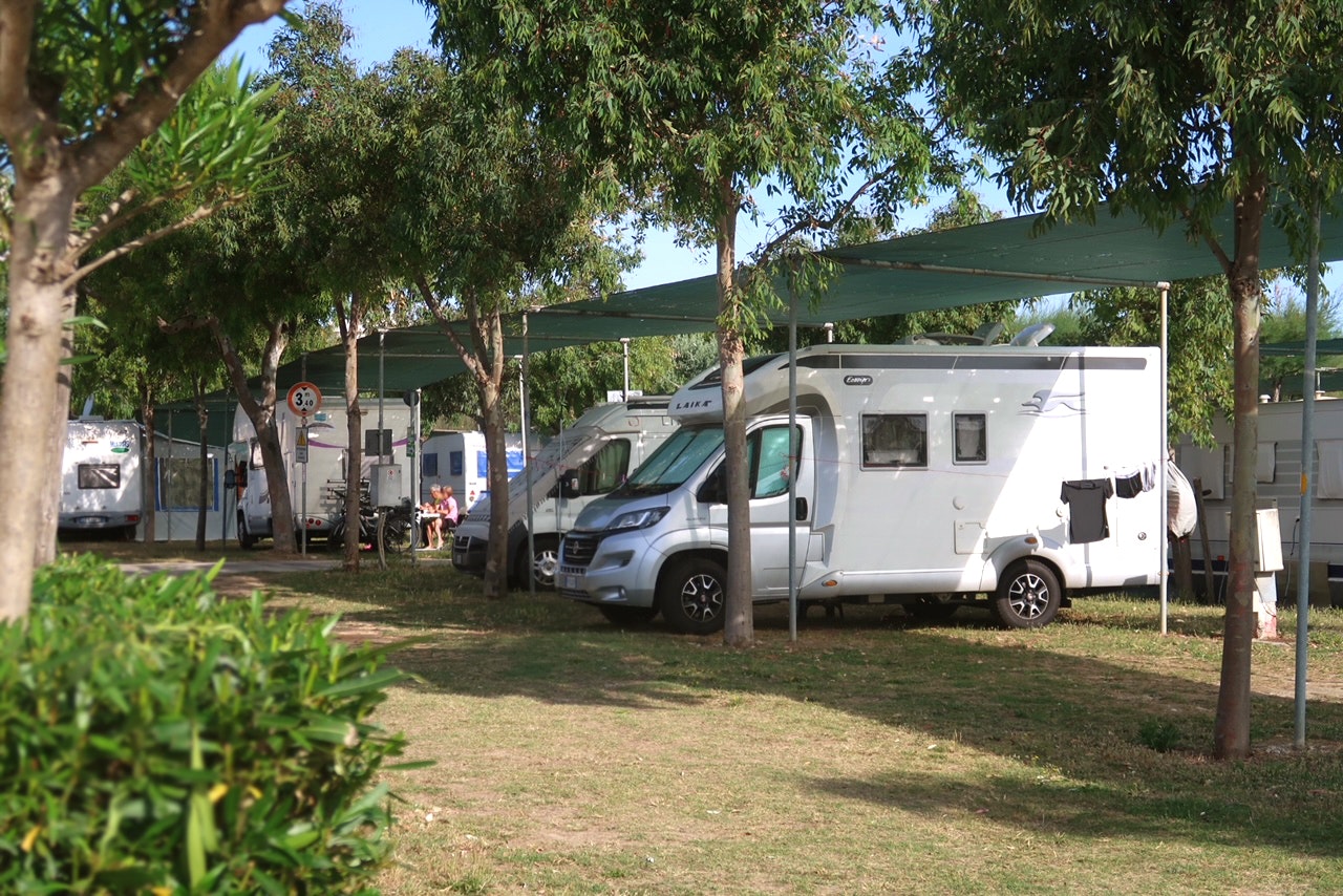 Wohnmobil unter einem Sonnensegel auf einem Campingplatz, daneben ein weiterer Camper und Bäume im Hintergrund.