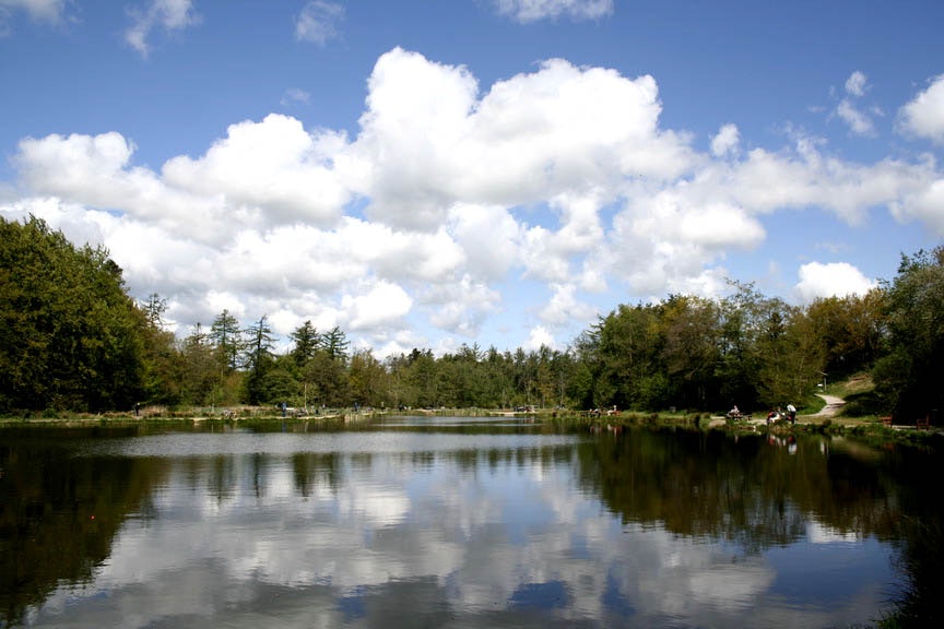 Ruhiger Campingsee mit Spiegelung im Wasser, umgeben von Wald und einem Weg am Ufer unter weißen Wolken.