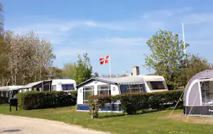 Wohnwagen auf einem Campingplatz mit Hecke, Rasen und Flagge, im Hintergrund weitere Wohnwagen und ein Zelt.