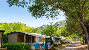 Reihe von Bungalows und Wohnwagen am Campingplatz, Sitzplätze mit Tischen, Bäume und Berge im Hintergrund.