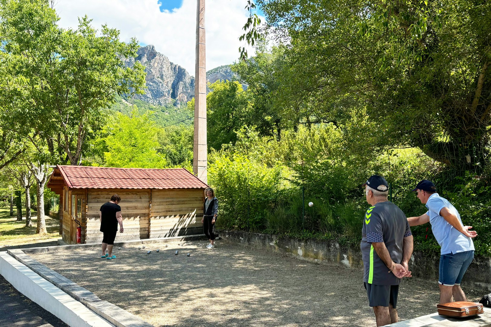 Weg auf dem Campingplatz zwischen Bäumen, Blick auf Berge, daneben ein kleines Gebäude mit Holzveranda.