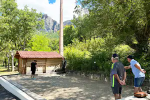 Weg auf dem Campingplatz zwischen Bäumen, Blick auf Berge, daneben ein kleines Gebäude mit Holzveranda.