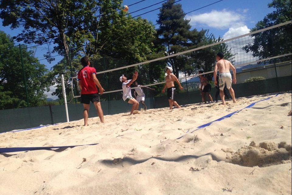 Beachvolleyballfeld mit Netz und Sand, mehrere Personen spielen Volleyball auf dem Campingplatz, im Hintergrund Bäume und Gebäude.