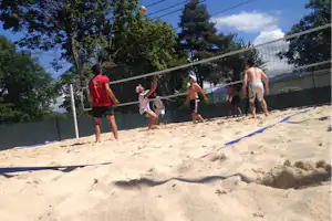 Beachvolleyballfeld mit Netz und Sand, mehrere Personen spielen Volleyball auf dem Campingplatz, im Hintergrund Bäume und Gebäude.