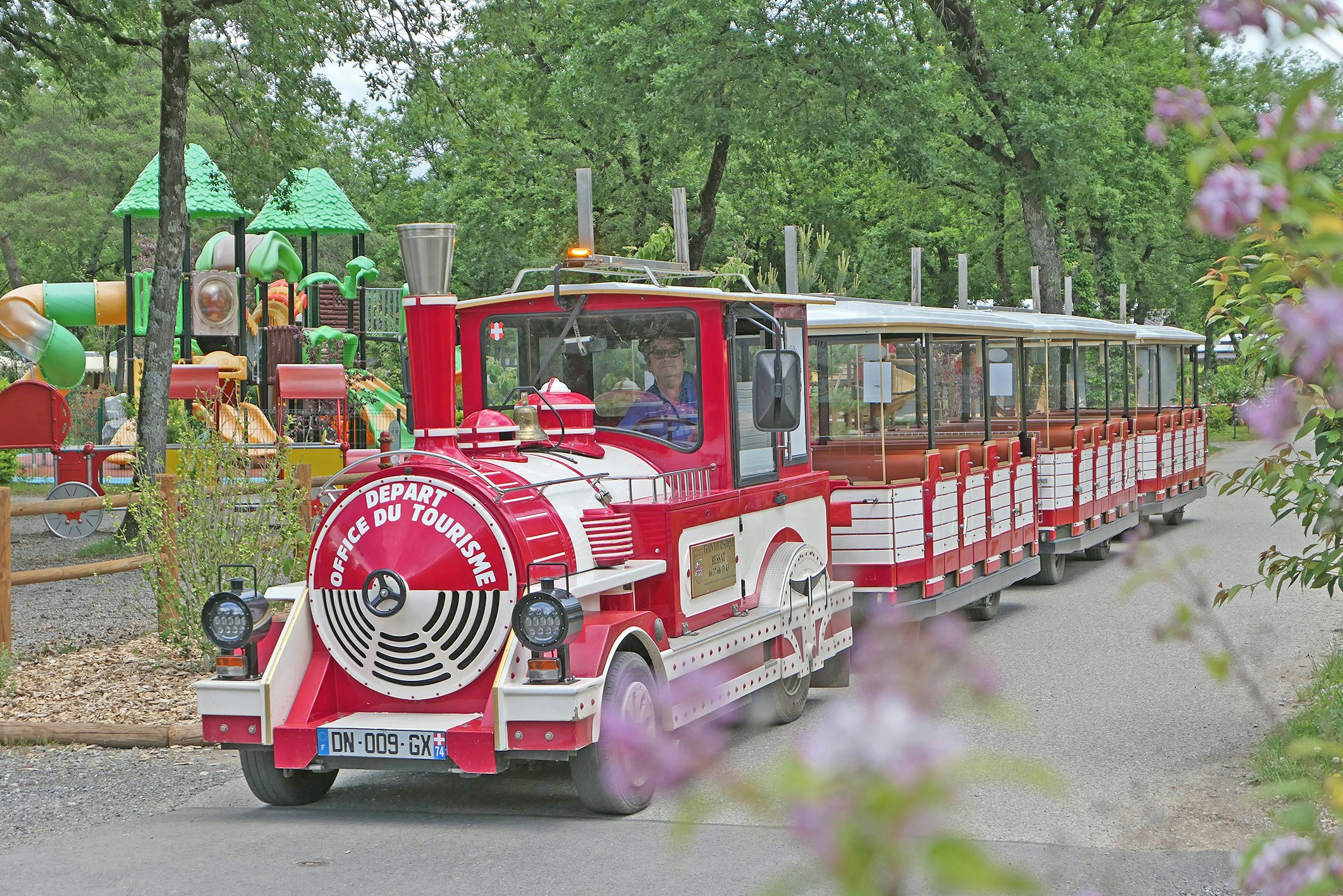 Roter kleiner Zug mit offenen Wagen auf einem Campingplatzweg, umgeben von Bäumen und grünen Spielgeräten.