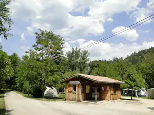 Holzgebäude am Campingplatz am Wegesrand, mit Eingang und Schildern, umgeben von Bäumen und Wiese.