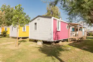 Ein Mobilheim mit Veranda und Holztreppe auf einem gepflegten Campingplatz, umgeben von Rasen und Bäumen.