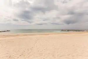 Breiter Sandstrand mit Blick auf das Meer, Wellen und eine Reihe Felsen am Ufer unter bewölktem Himmel.