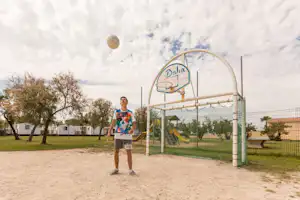 Ein Mann steht auf einem Campingplatz vor einem Basketballkorb auf einem Sandplatz, im Hintergrund Bäume und Wohnwagen.