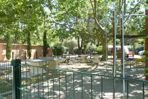 Spielplatz mit grünem Zaun, Schaukelgestell und zwei weißen Picknicktischen unter großen Bäumen im Campingbereich.