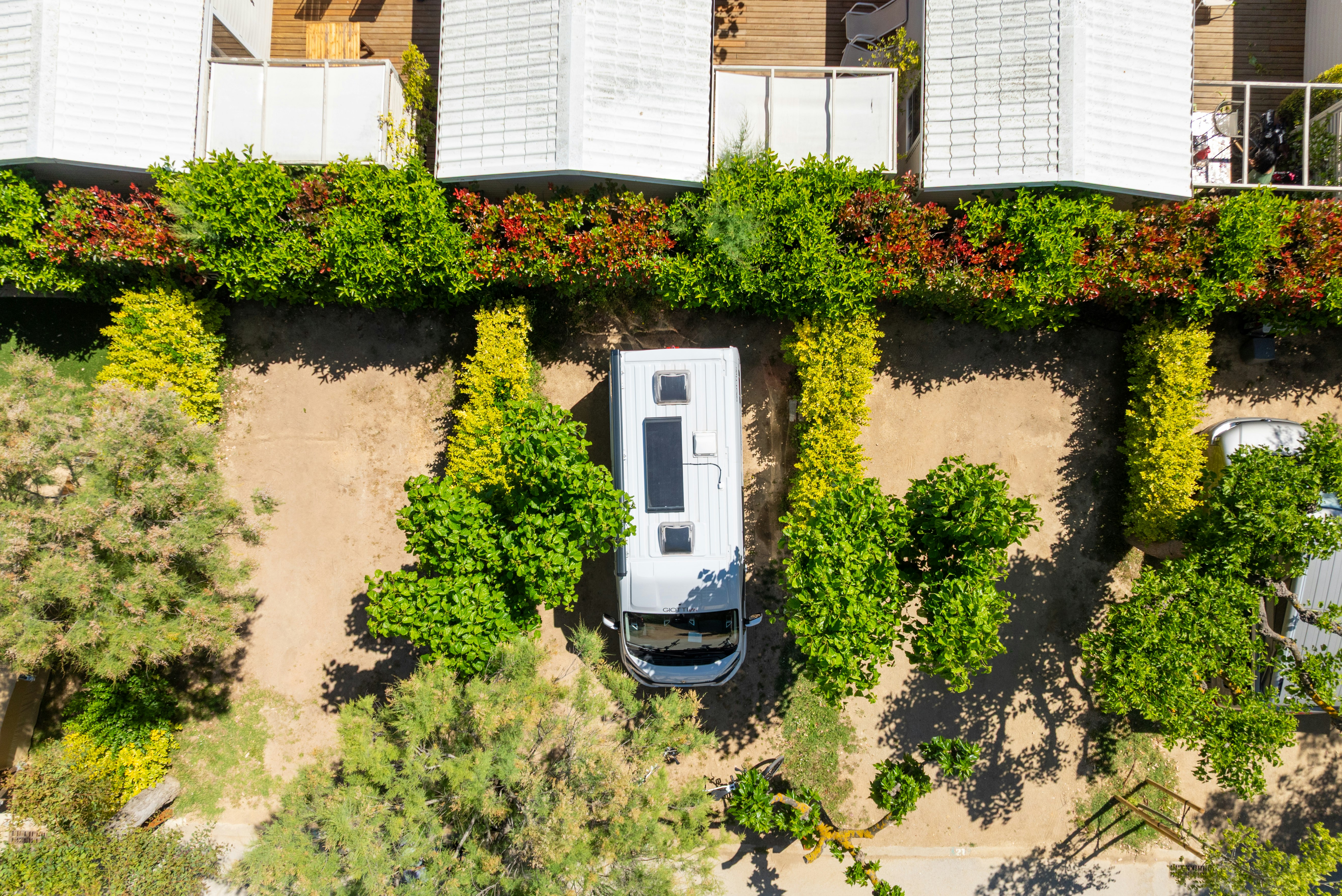 Wohnmobil auf einem Stellplatz zwischen Hecken, mit gepflastertem Weg und weißen Gebäuden im Hintergrund.