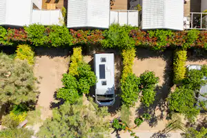 Wohnmobil auf einem Stellplatz zwischen Hecken, mit gepflastertem Weg und weißen Gebäuden im Hintergrund.