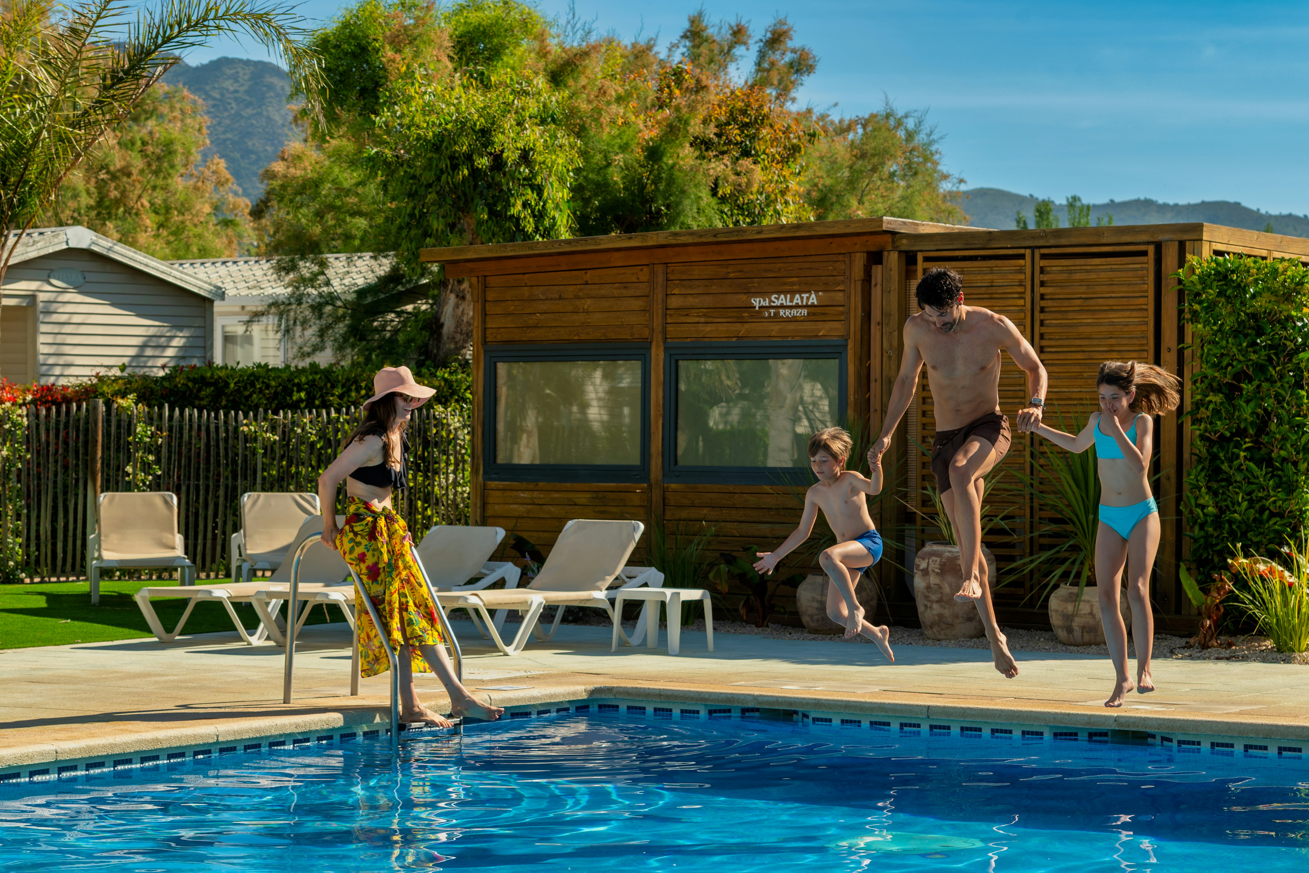 Familie am Campingpool mit Sonnenliegen vor einem Holzgebäude, im Hintergrund Bäume und Berge.