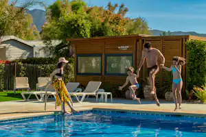 Familie am Campingpool mit Sonnenliegen vor einem Holzgebäude, im Hintergrund Bäume und Berge.