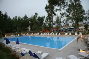 Campingpool mit blauem Wasser, weißen Liegen und Sonnenschirmen, umgeben von Bäumen und beleuchteten Wegen.