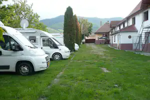 Parkende Autos auf einem Campingplatz, Rasenweg und Wohngebäude mit Terrasse, im Hintergrund Berge und Bäume.