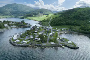 Campingplatz am Fluss mit Wohnwagen auf Inseln, Stegen und Wegen, umgeben von grünen Bergen und Tälern.