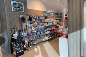 Camping-Shop mit Regalen voller Campingzubehör und Produkten, beleuchteter Verkaufsfläche und Eingang zwischen Holzwänden.