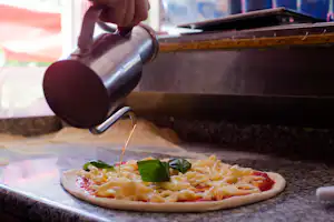 Pizza auf einer Grillplatte im Campingbereich, daneben eine Person mit Getränkekanne und Kräutern auf dem Belag.