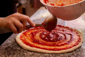 Hand legt Tomatensauce auf eine Pizza auf einer Arbeitsfläche, daneben Schüssel mit Sauce, beim Camping.