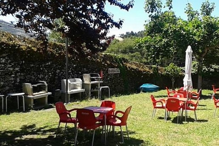 Camping-Garten mit roten Stühlen und Tischen, Sonnenschirm und Liegestühlen am Rasen, umgeben von Hecken und Bäumen.