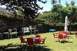 Camping-Garten mit roten Stühlen und Tischen, Sonnenschirm und Liegestühlen am Rasen, umgeben von Hecken und Bäumen.