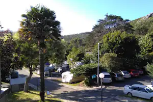 Campingplatz mit Baumreihe, Wohnwagen und Zelten auf Stellplätzen, Parkplatz mit Autos und Blick in die grüne Hügellandschaft.