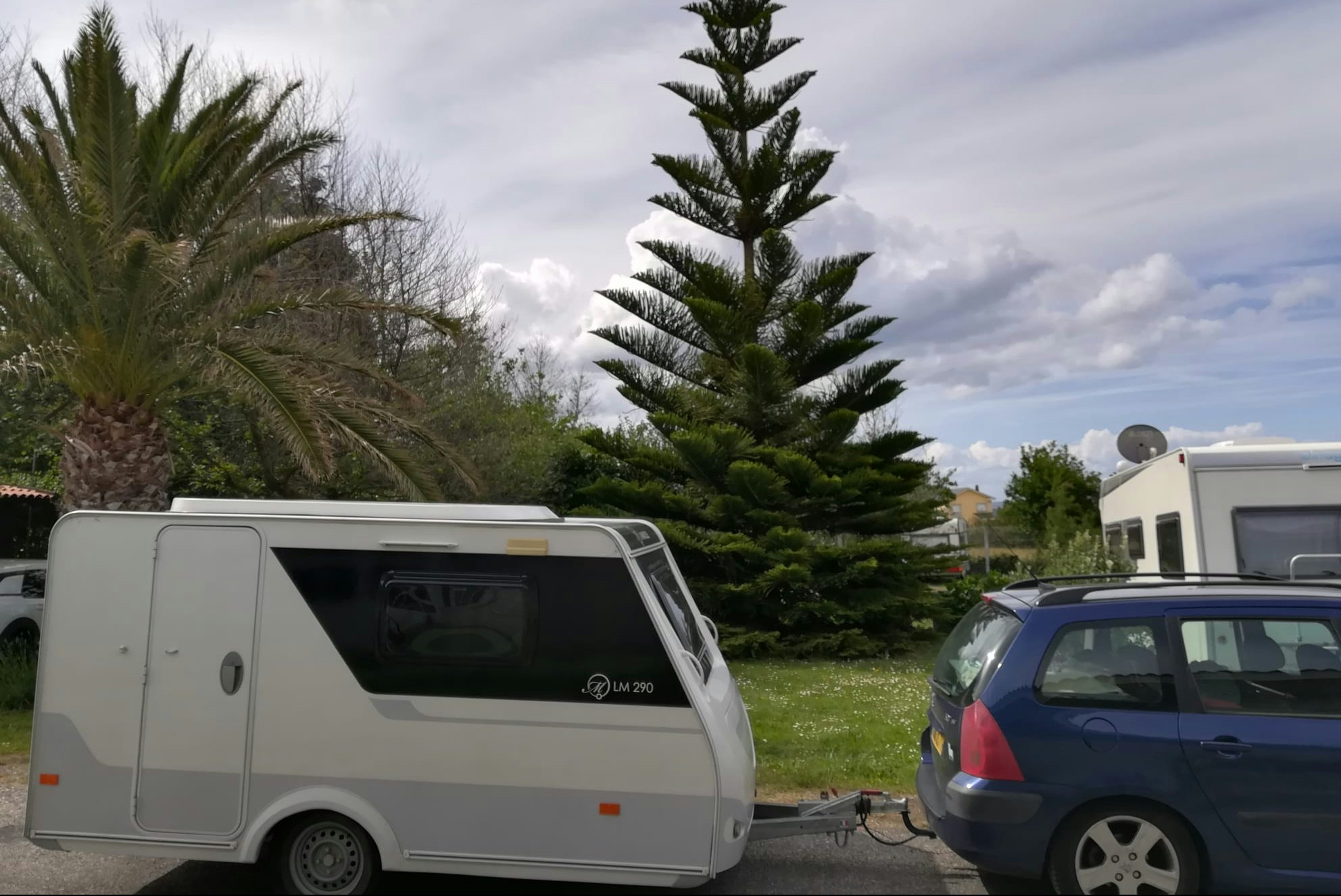 Wohnwagen und Auto auf dem Stellplatz, im Hintergrund Palmen und ein gepflegter Garten mit Rasenfläche.