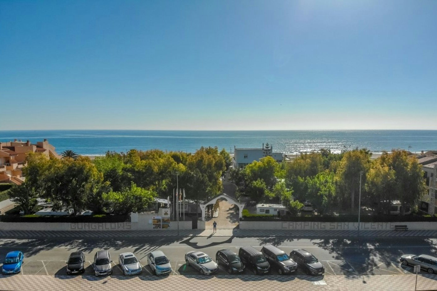 Parkplatz mit mehreren Autos und Blick auf das Meer, dahinter Bäume und Häuser entlang einer Küstenstraße.
