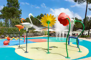Wasserspiel am Pool mit bunten Figuren und Sprühdüsen, im Hintergrund Liegen und Bäume vor blauem Himmel.