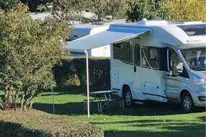 Weißes Wohnmobil auf einem Campingplatz mit Markise, Tisch und Stühlen am Stellplatz, umgeben von Bäumen und Rasen.
