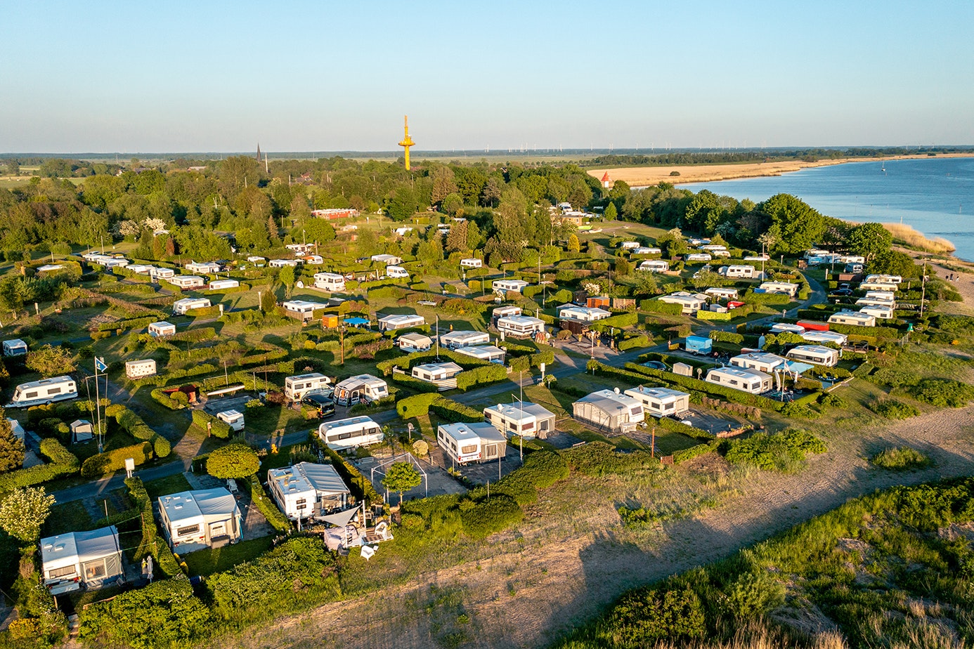 Luftaufnahme eines Campingplatzes mit Wohnwagen und Zelten, Wegen und Grünflächen, am Flussufer mit Bäumen und Häusern in der Ferne.