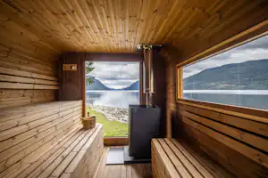 Holzsauna innen mit Bank und Ofen, große Fenster mit Blick auf See und Berge, Holzdecke und Wände.