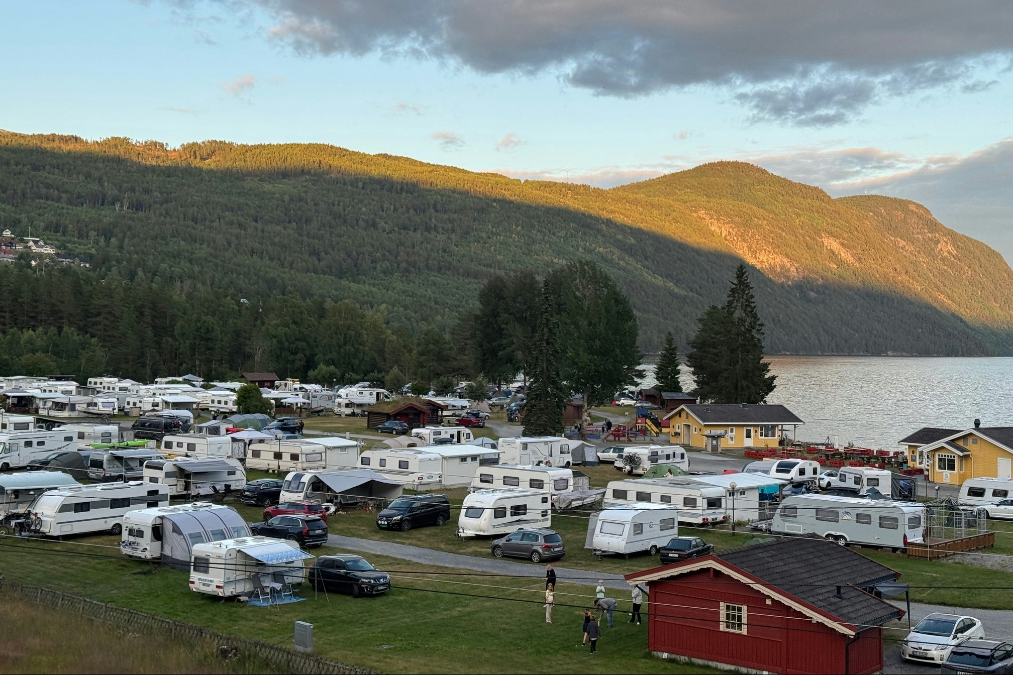 Wohnmobile und Campingplätze am Hang, mit roten Gebäuden, Bäumen und Blick auf einen See zwischen bewaldeten Hügeln.