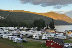 Wohnmobile und Campingplätze am Hang, mit roten Gebäuden, Bäumen und Blick auf einen See zwischen bewaldeten Hügeln.