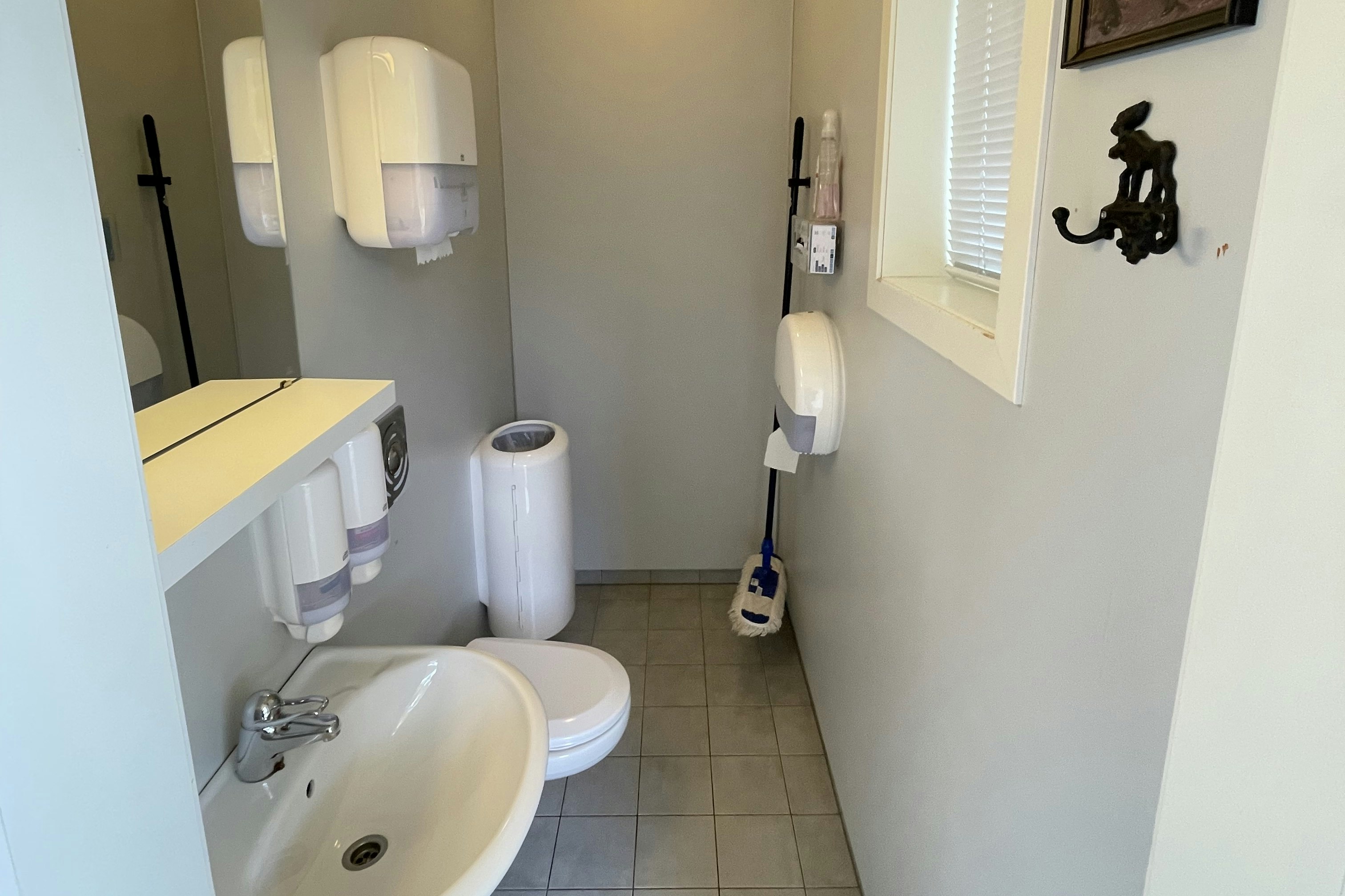 Helles Badezimmer mit Waschbecken, Toilette und Duschbereich, Fliesenboden und Fenster mit Jalousie.