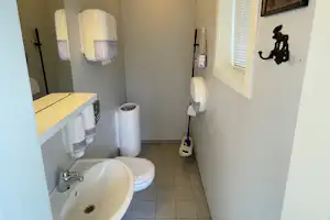 Helles Badezimmer mit Waschbecken, Toilette und Duschbereich, Fliesenboden und Fenster mit Jalousie.