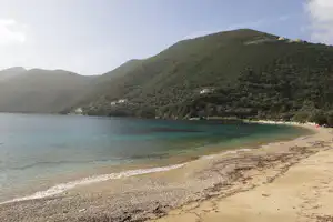 Blick auf einen Sandstrand an einer Bucht mit türkisfarbenem Wasser und bewaldeten Hügeln im Hintergrund.