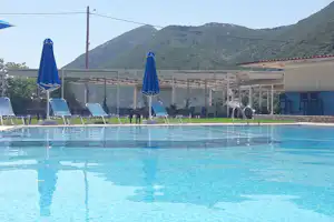 Campingpool mit blauen Sonnenschirmen und Liegestühlen, im Hintergrund ein überdachter Bereich und grüne Hügel.
