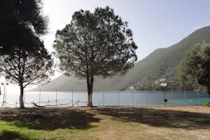 Campingplatz am See mit Grasfläche, zwei Bäumen und Hängematte am Ufer, im Hintergrund Berge und ruhiges Wasser.