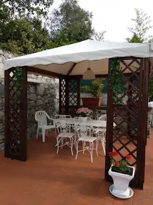 Camping-Terrasse mit weißem Sonnendach, Holzgitter, weißen Metallstühlen und Tisch vor Steinmauer, umgeben von Büschen und Pflanzen.