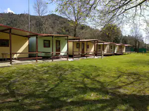 Reihe von Bungalows mit Holzterrassen, grüner Wiese und Bäumen im Hintergrund vor einem bewaldeten Hügel.