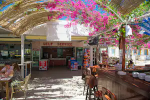 Self-Service-Kiosk mit Theke und Kühlboxen unter einem Blumenbogen mit pinken Blüten, daneben Tische und Stühle.
